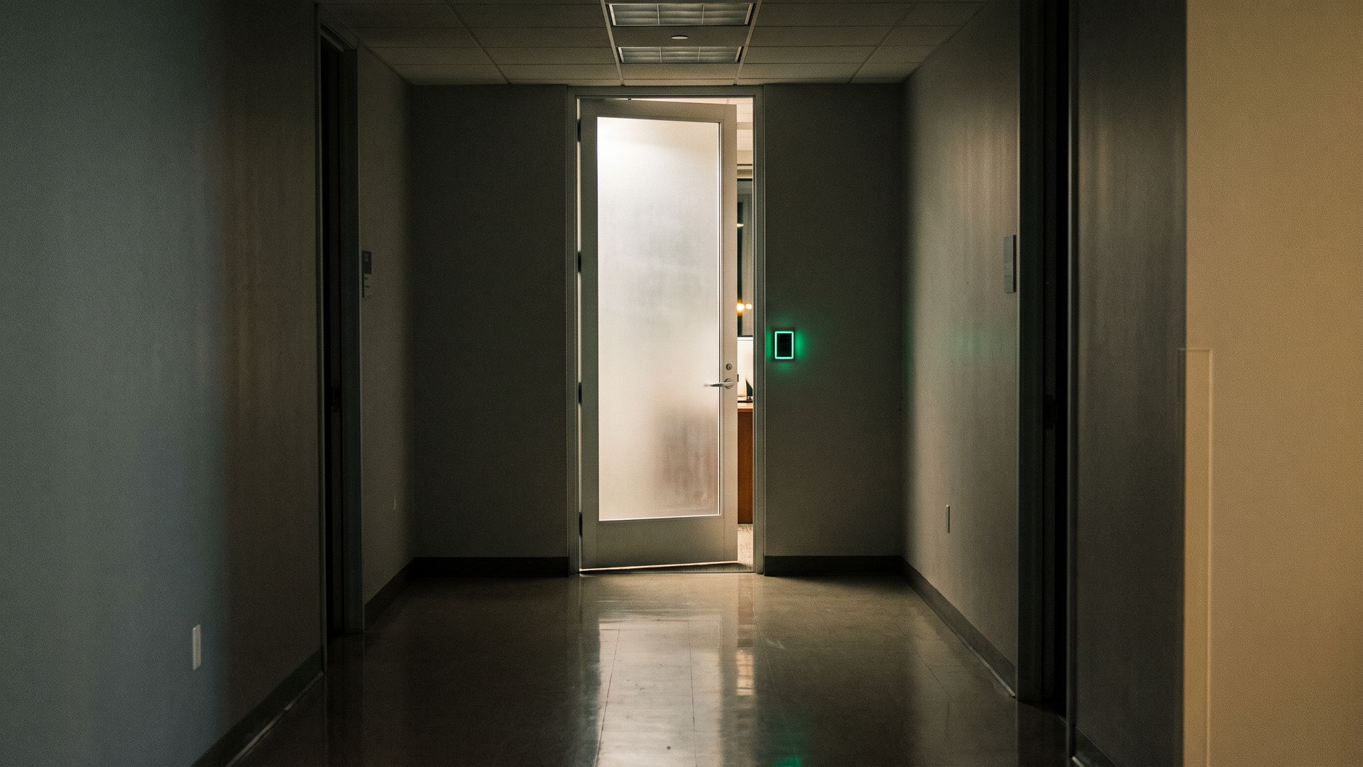 Una porta a vetri socchiusa in un corridoio d'ufficio buio, con un lettore di tessere appena acceso
