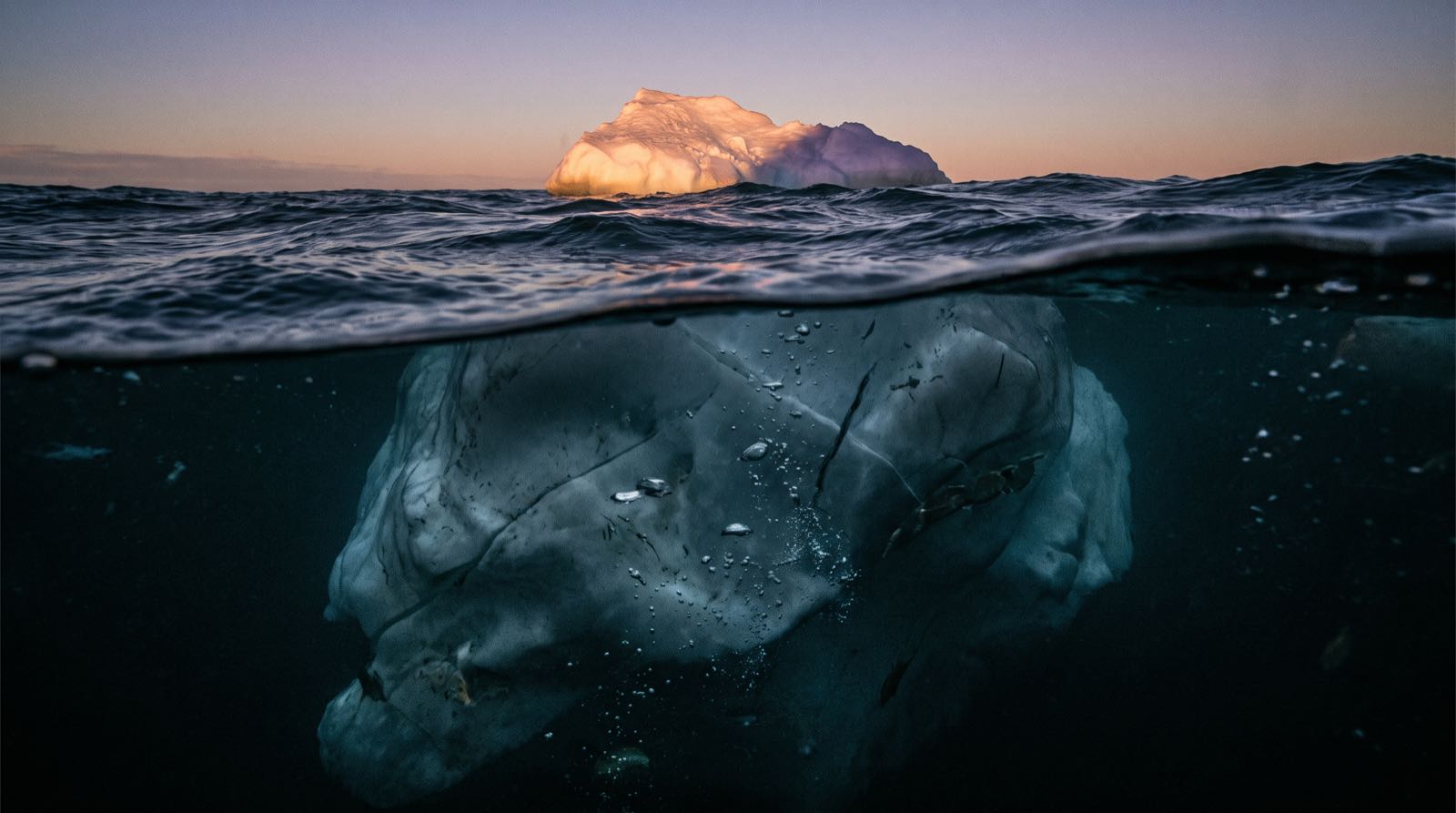 Un iceberg fotografato a metà tra sopra e sotto il pelo dell'acqua: una piccola punta illuminata dal tramonto emerge, mentre una massa scura enorme incombe sott'acqua