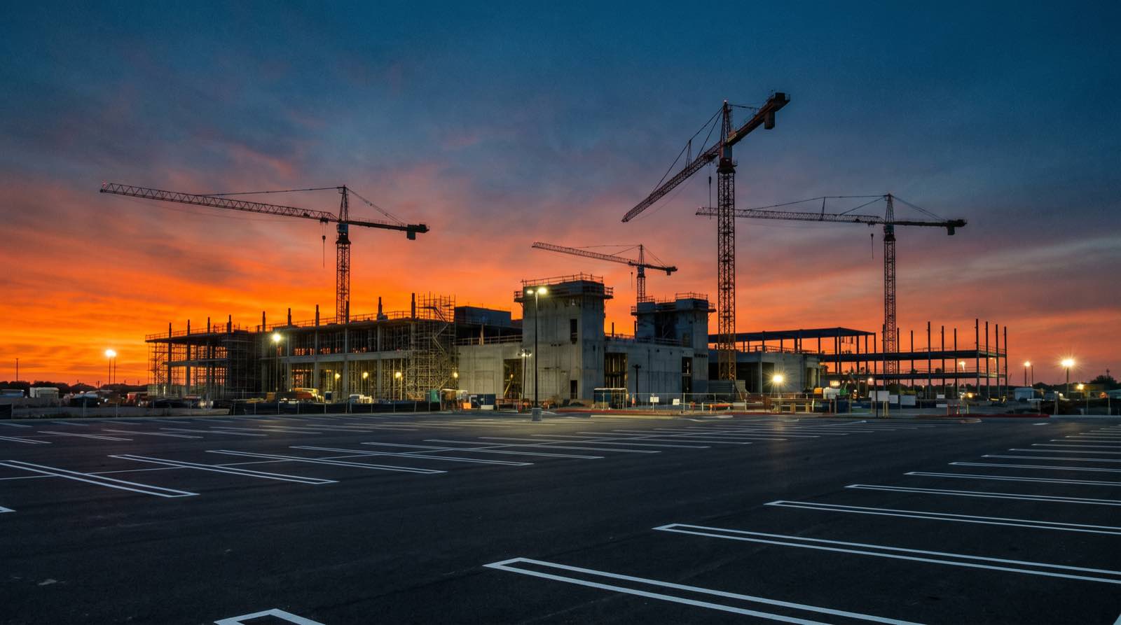 Un enorme stabilimento per semiconduttori vuoto e ancora in costruzione al tramonto, gru silhouette contro un cielo arancione, un parcheggio vuoto in primo piano